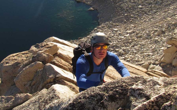 A person looks up at the camera while rock climbing on an outward bound gap year course. There is a vast rocky landscape and an alpine lake below them. 